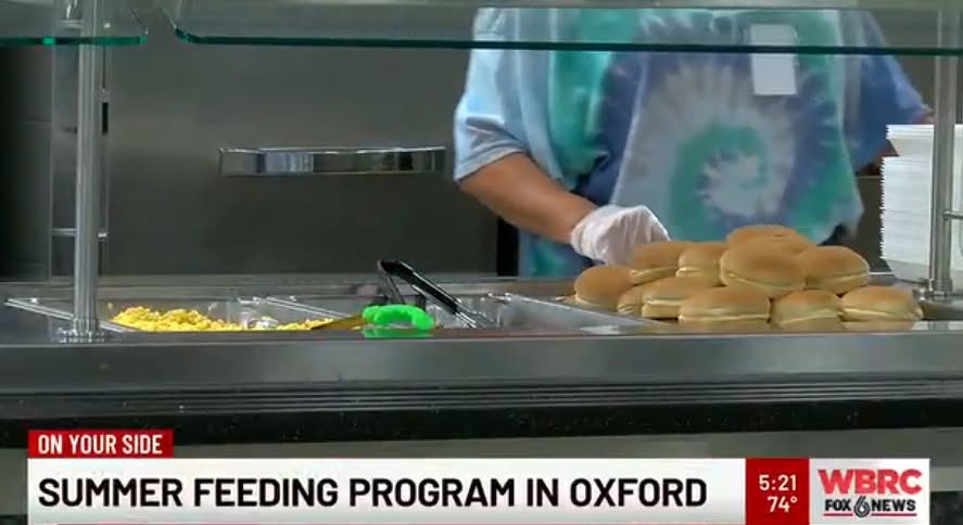 WBRC Fox 6 News broadcast still captioned Summer Feeding Program in Oxford, showing a school nutrition worker in gloves plating hamburger buns and corn on a cafeteria serving line