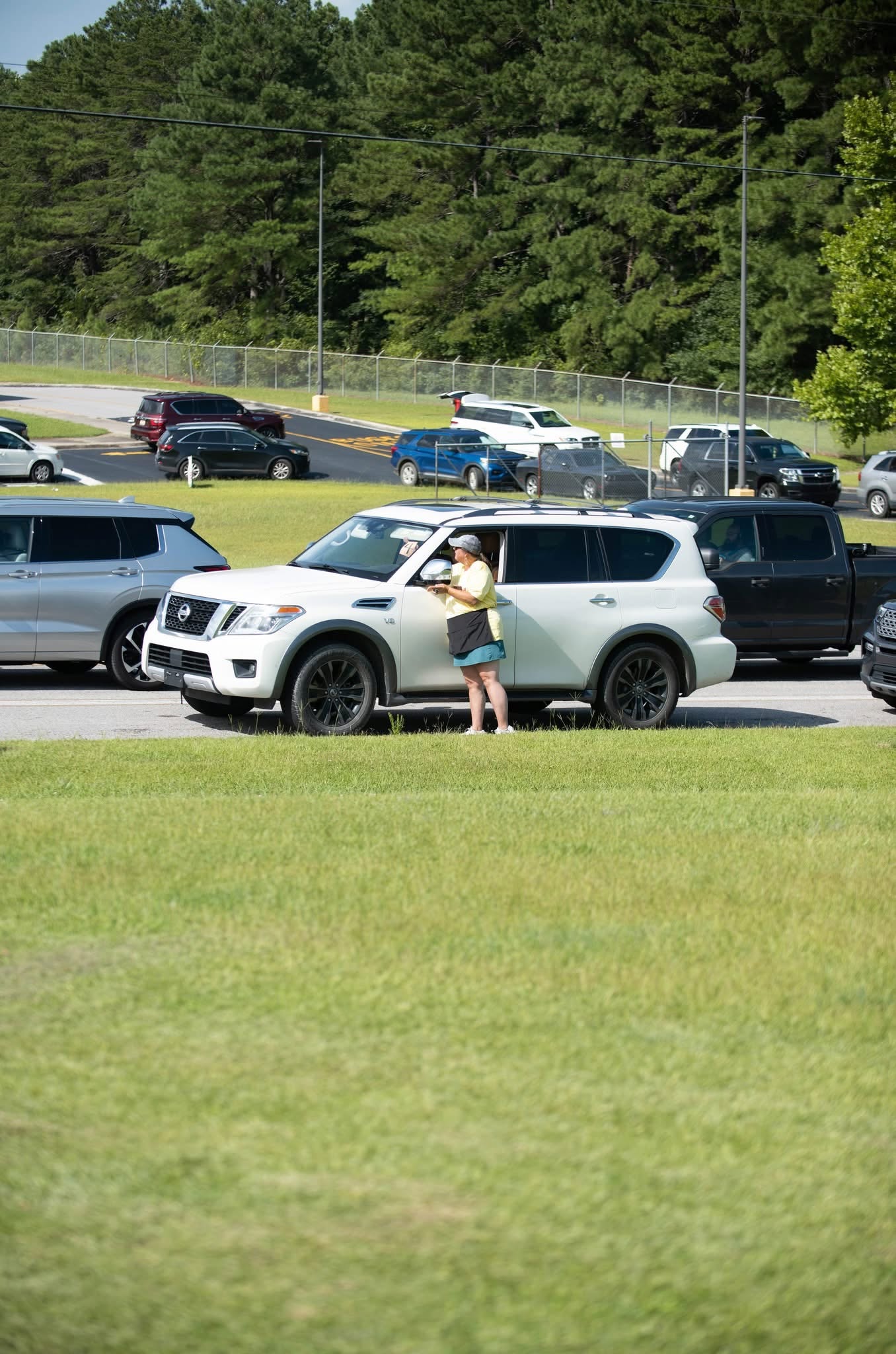 Staff member serving a family through a car window during a curbside summer feeding distribution, with a line of vehicles waiting